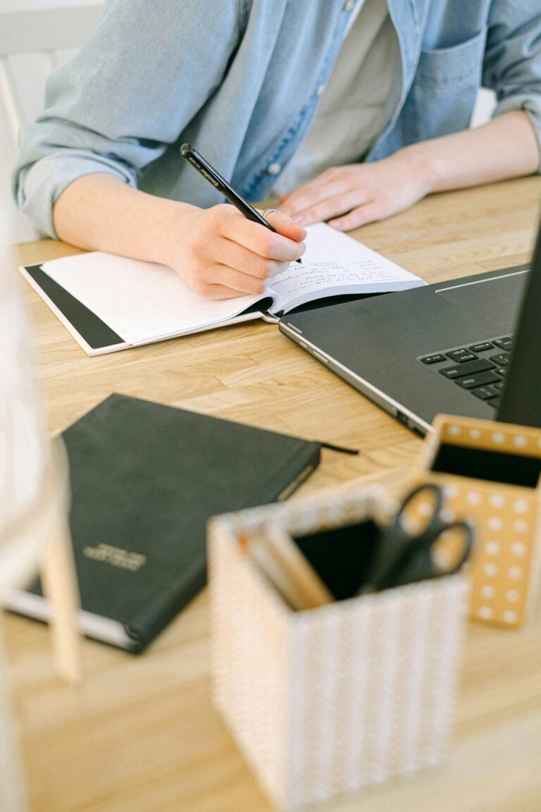 Adult writing notes on a notebook while working on a laptop in a cozy home office environment.